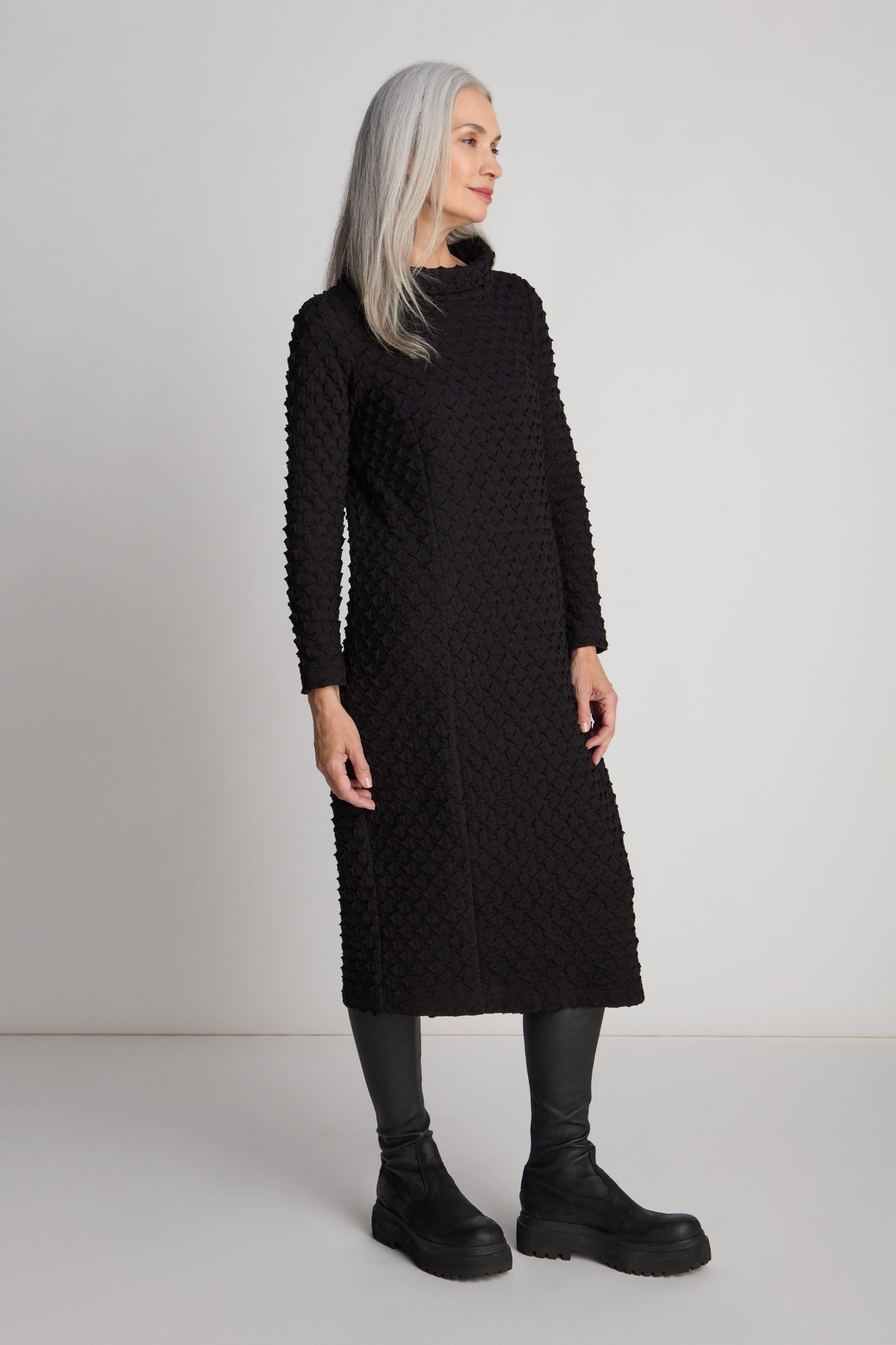 A woman with long gray hair is standing and wearing the Pocket Jersey Cowl Neck Dress, which showcases a flattering slim silhouette paired with black leggings and boots. She looks to her right against a plain light background.