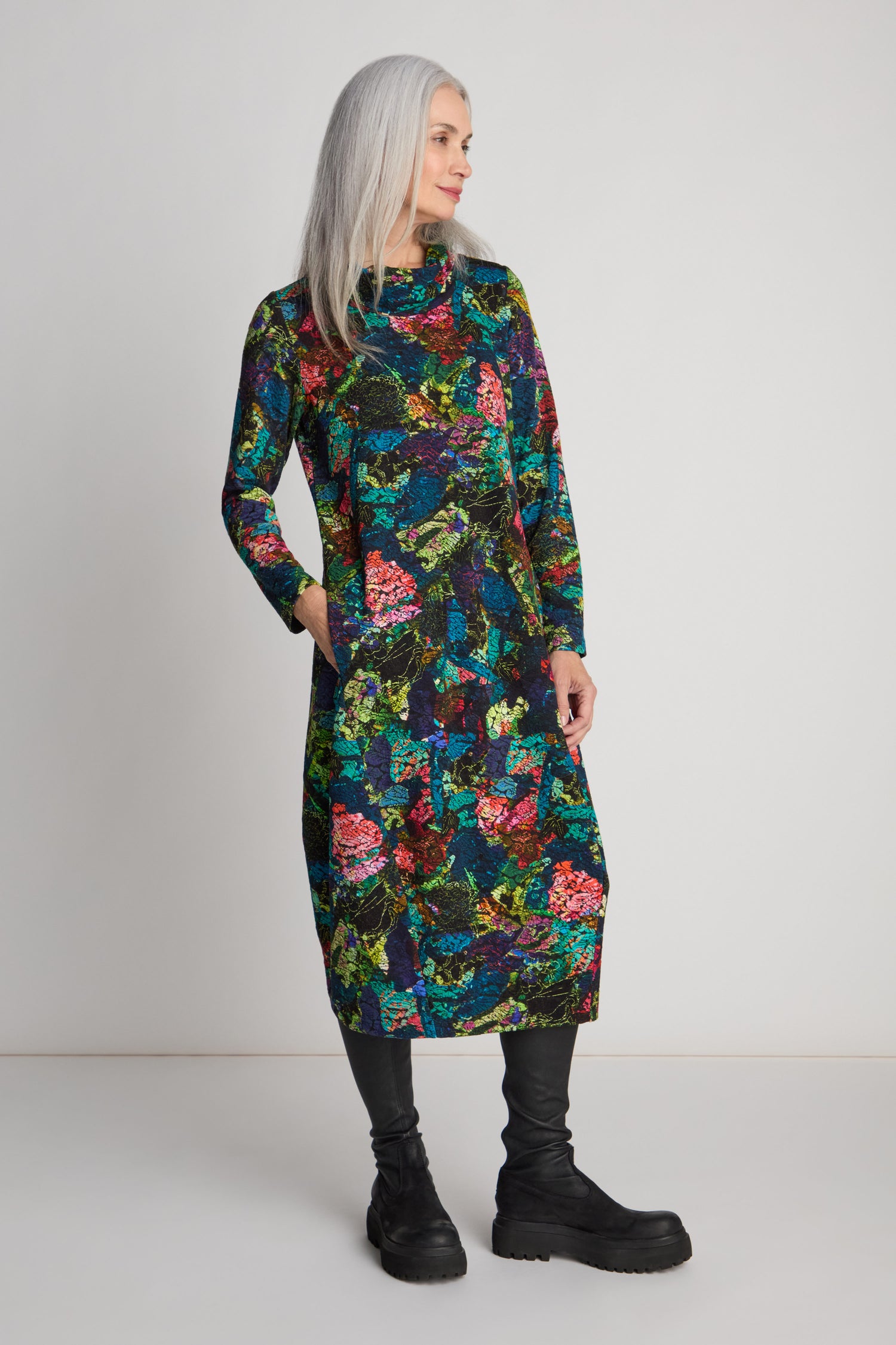 A person with long gray hair, dressed in an Abstract Floral Jersey Bubble Dress featuring pockets, stands against a plain background, gazing to the side. They are also wearing black leggings and black boots.