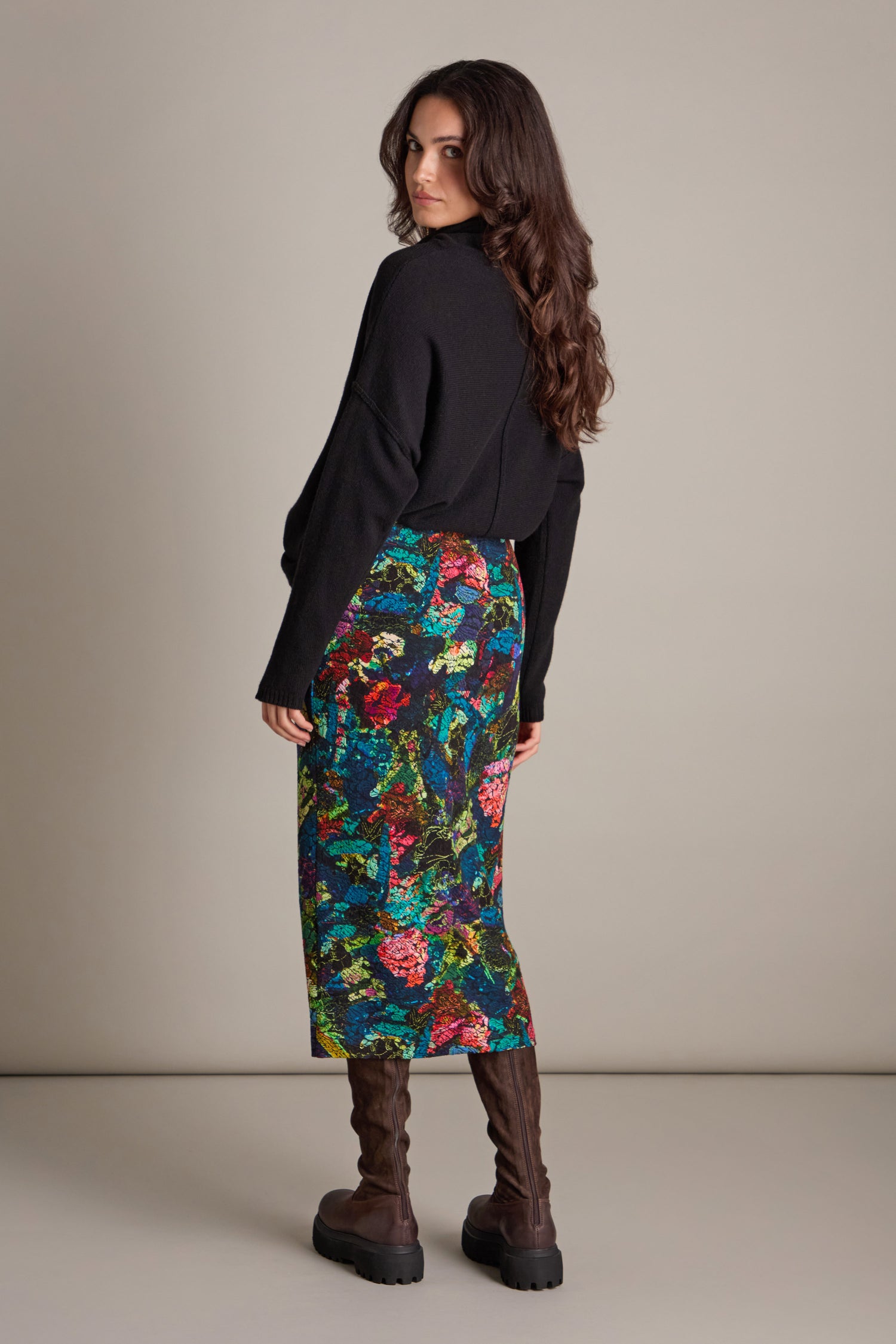 A person with long hair, wearing a black sweater and the Abstract Floral Jersey Tube Skirt that radiates artistic elegance, stands against a plain background, looking back over their shoulder in stylish brown boots.