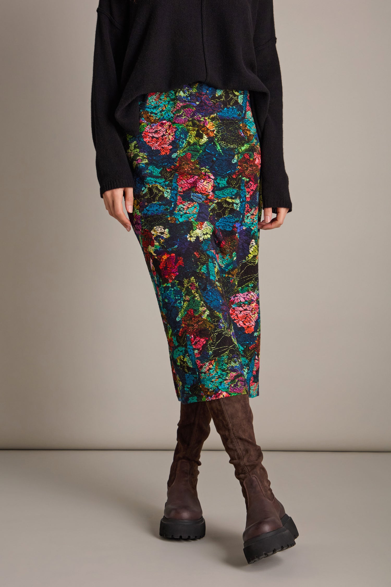 A person wearing a black sweater and the Abstract Floral Jersey Tube Skirt, paired with brown knee-high boots, stands against a plain background.