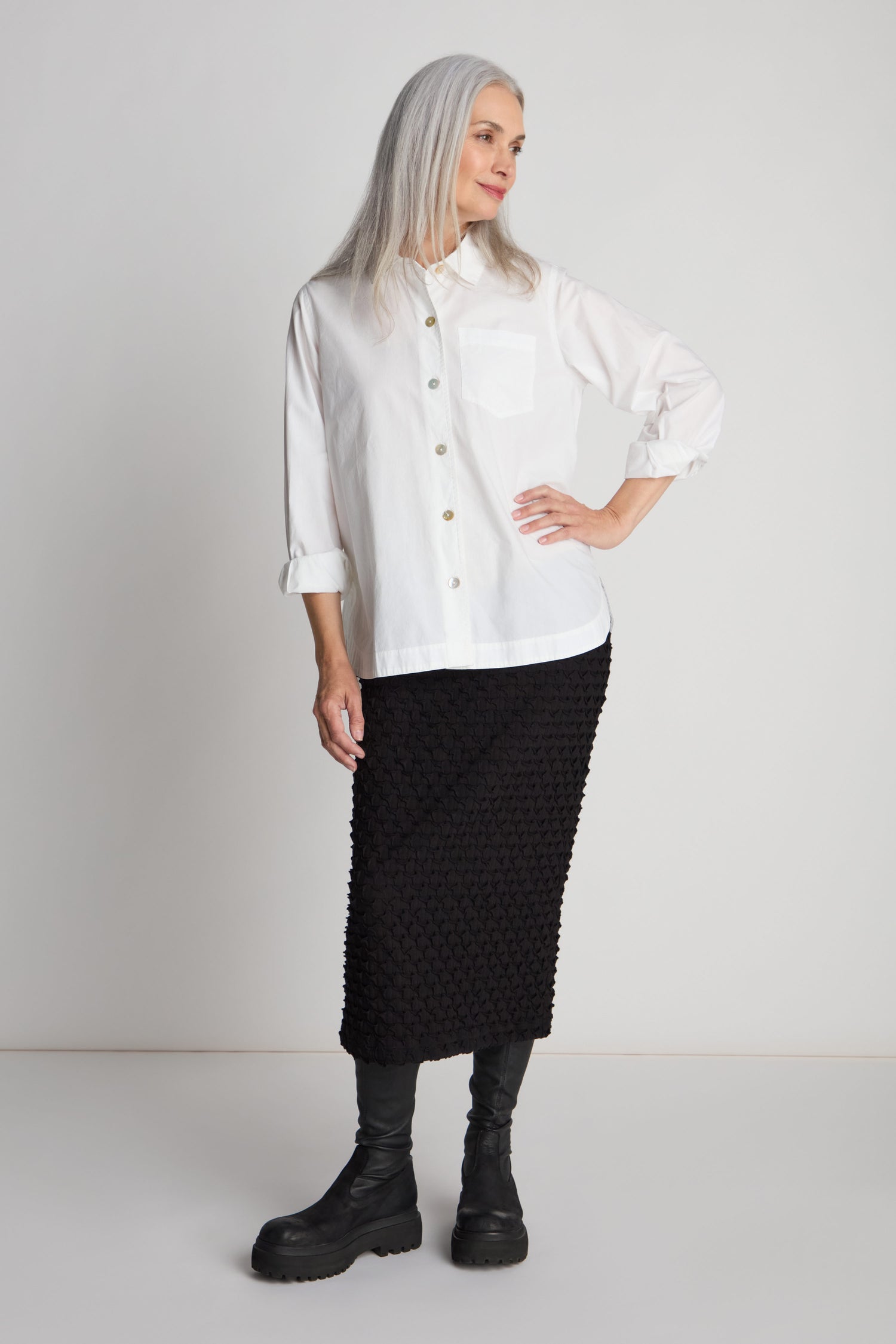 A woman with long, gray hair wearing a white button-up shirt and the Pocket Jersey Tube Skirt in black, which features a textured fabric and an elasticated waist, stands against a plain background. She has one hand on her hip and looks off to the side.