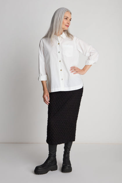 A woman with long, gray hair wearing a white button-up shirt and the Pocket Jersey Tube Skirt in black, which features a textured fabric and an elasticated waist, stands against a plain background. She has one hand on her hip and looks off to the side.