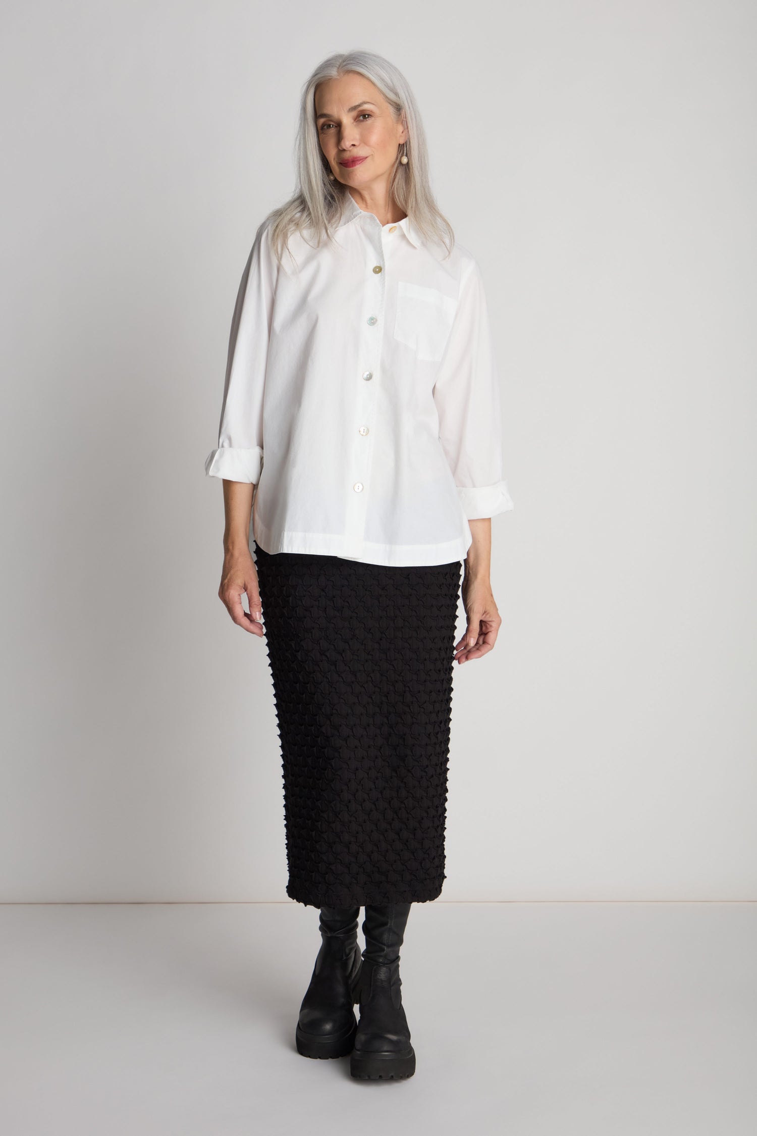 A woman with long gray hair wearing a white button-up shirt and the Pocket Jersey Tube Skirt, which features an elasticated waist, stands in front of a plain backdrop, looking forward. She is also wearing black ankle boots.