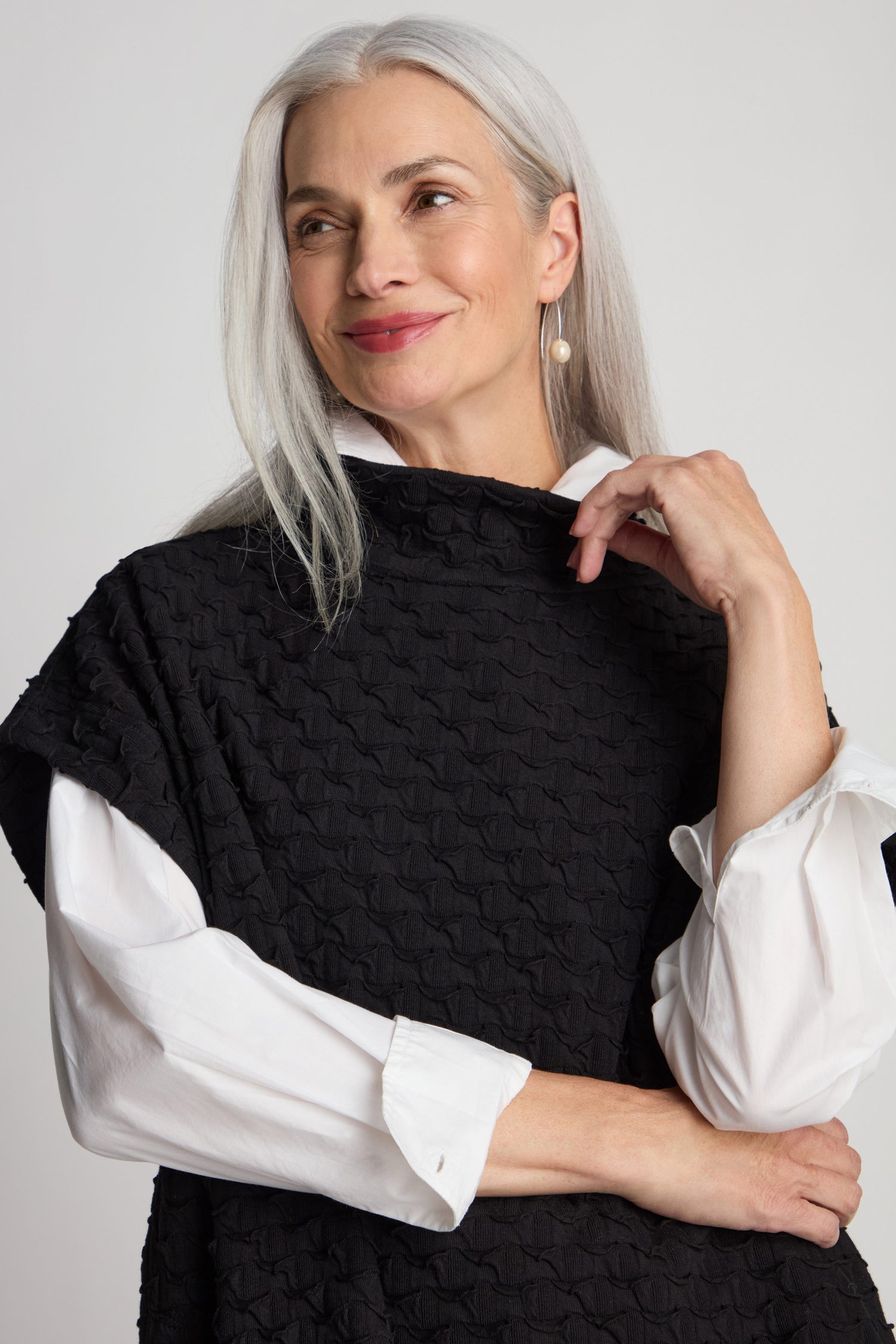 A woman with long gray hair smiles while wearing a Pocket Jersey Tabard over a white shirt featuring a luxurious texture. She holds the collar of her tabard with her left hand.