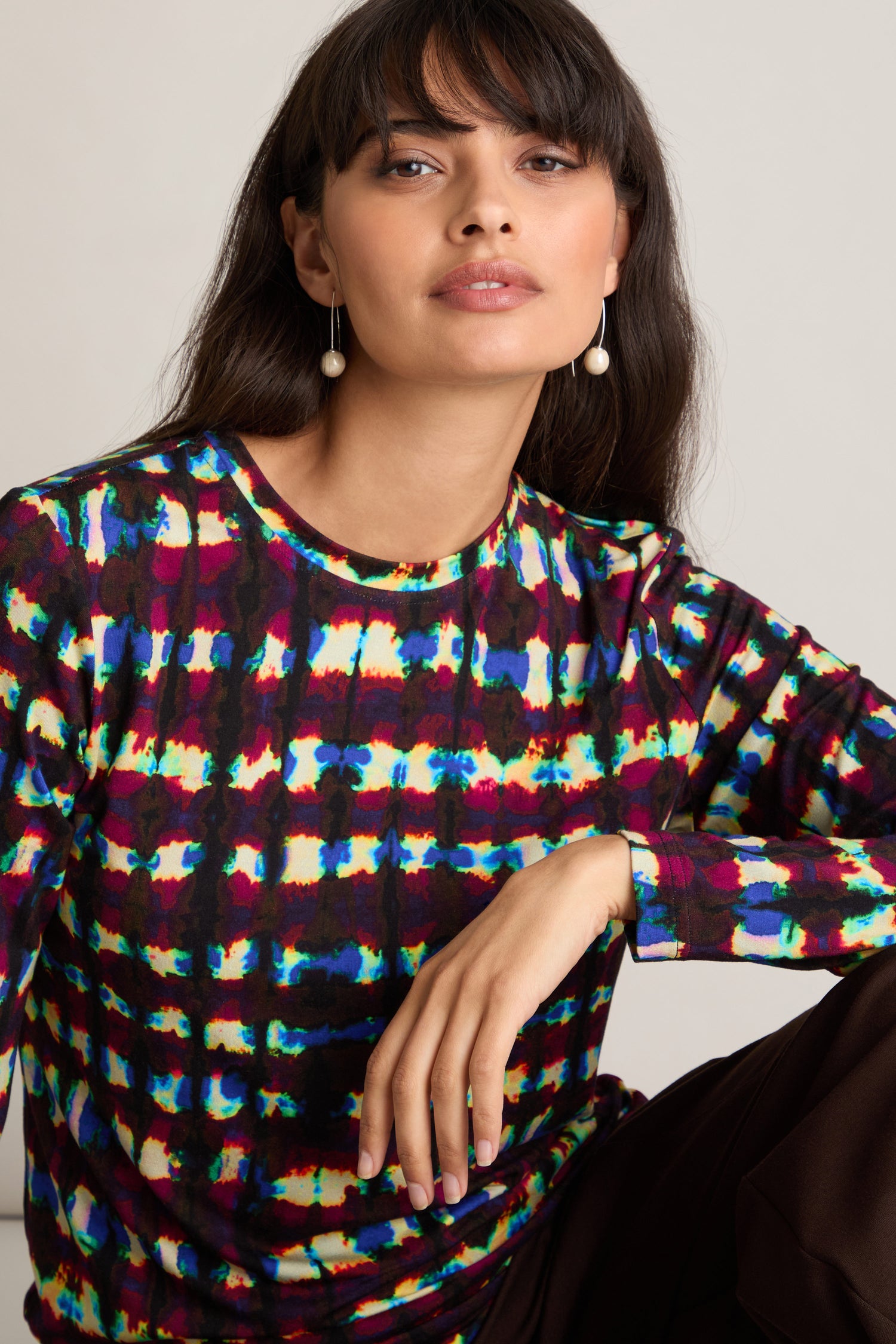 A woman with long dark hair is wearing pearl earrings and a colorful Marble Stripe Jersey Top, posing with her hand resting on her knee against a plain background.