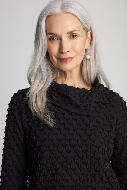 A woman with long white hair, wearing the Pocket Jersey Collar Top with an asymmetric collar and textured black design, and pearl earrings smiles at the camera.