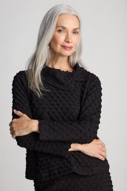 A woman with long gray hair, wearing the Pocket Jersey Collar Top in black with its unique textured design and asymmetric collar, stands with her arms crossed and smiles slightly against a plain background.