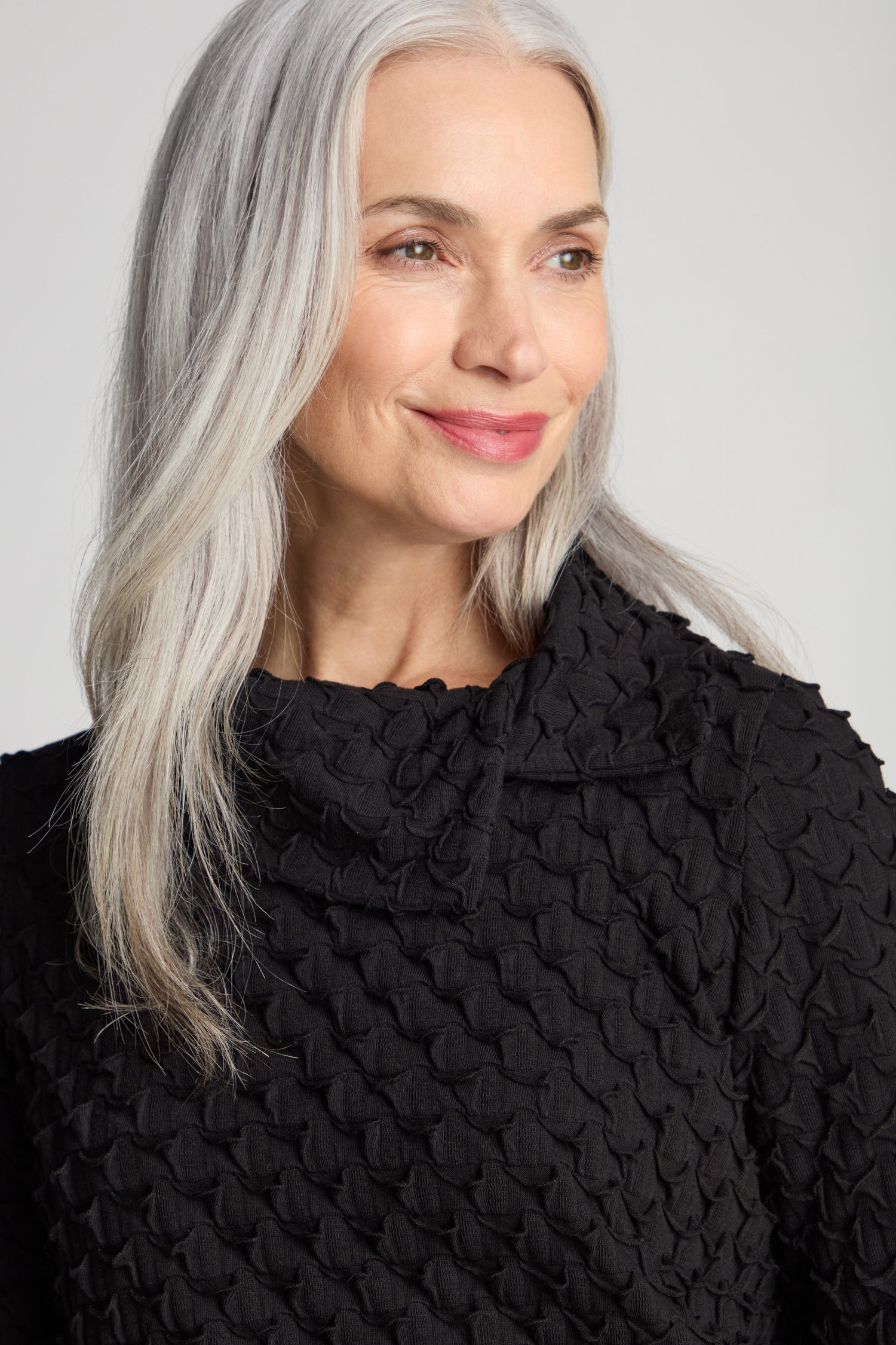 A smiling woman with long gray hair, wearing the Pocket Jersey Collar Top with its distinctive textured fabric and asymmetric collar, looks slightly to the side against a plain background.