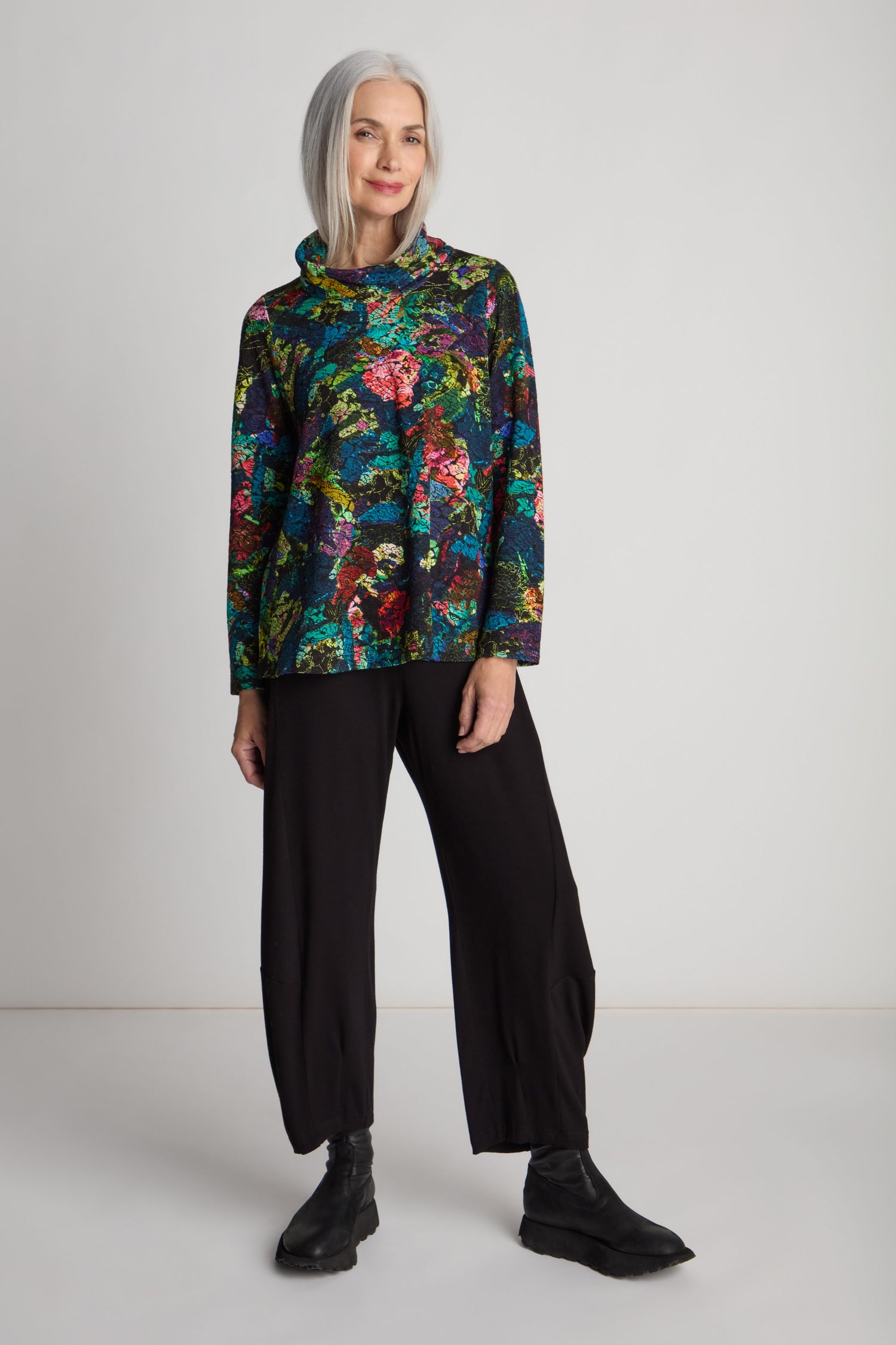 A woman with gray hair is wearing an "Abstract Floral Jersey Cowl Neck Top" with a flared silhouette, paired with black pants and black boots. She stands against a plain white background.