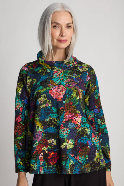 A woman with long, gray hair wears the Abstract Floral Jersey Cowl Neck Top with long sleeves. She stands against a plain background, exuding effortless elegance.