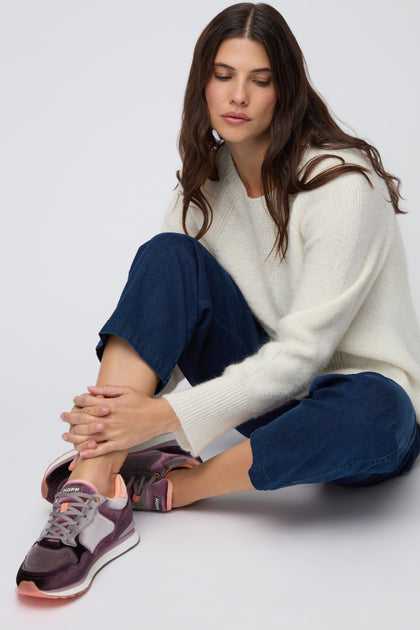 A woman with long brown hair, dressed in a white sweater, blue jeans, and Purple City Velvet Trainers, sits cross-legged on the floor, looking down.