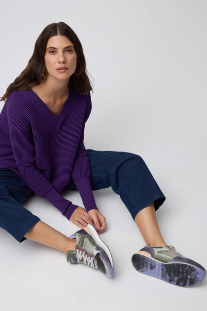 A woman with long hair sits on the floor against a plain light background, wearing a purple V-neck sweater, cropped blue jeans, and Green City Velvet Trainers.