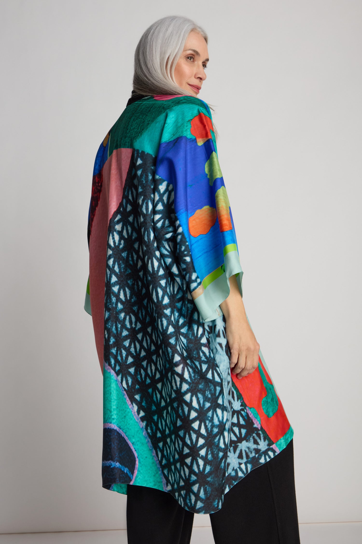 A person with long gray hair wears a colorful, patterned Ibiza Silk Kimono by Isabelle Gougenheim paired with black pants, glancing over their shoulder.