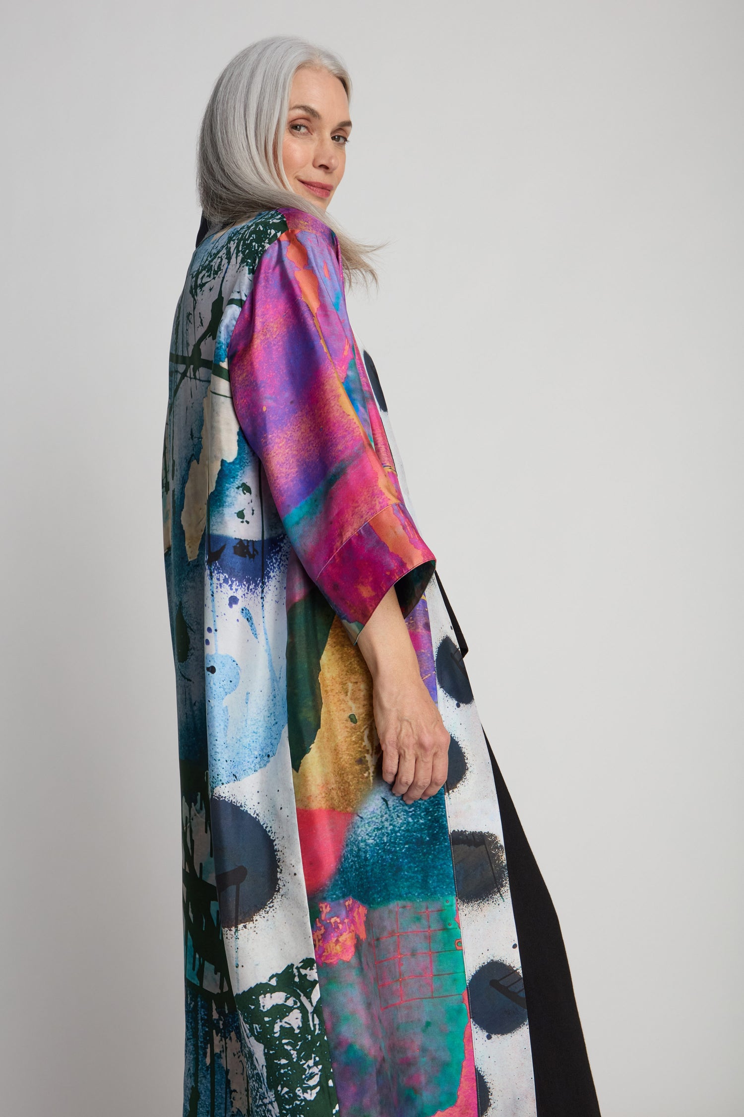 A woman with long gray hair wears a colorful, abstract-patterned coat by Isabelle Gougenheim. She is turned slightly to the side and looks over her shoulder, revealing a glimpse of the luxurious Connect the Dots Long Silk Kimono underneath.