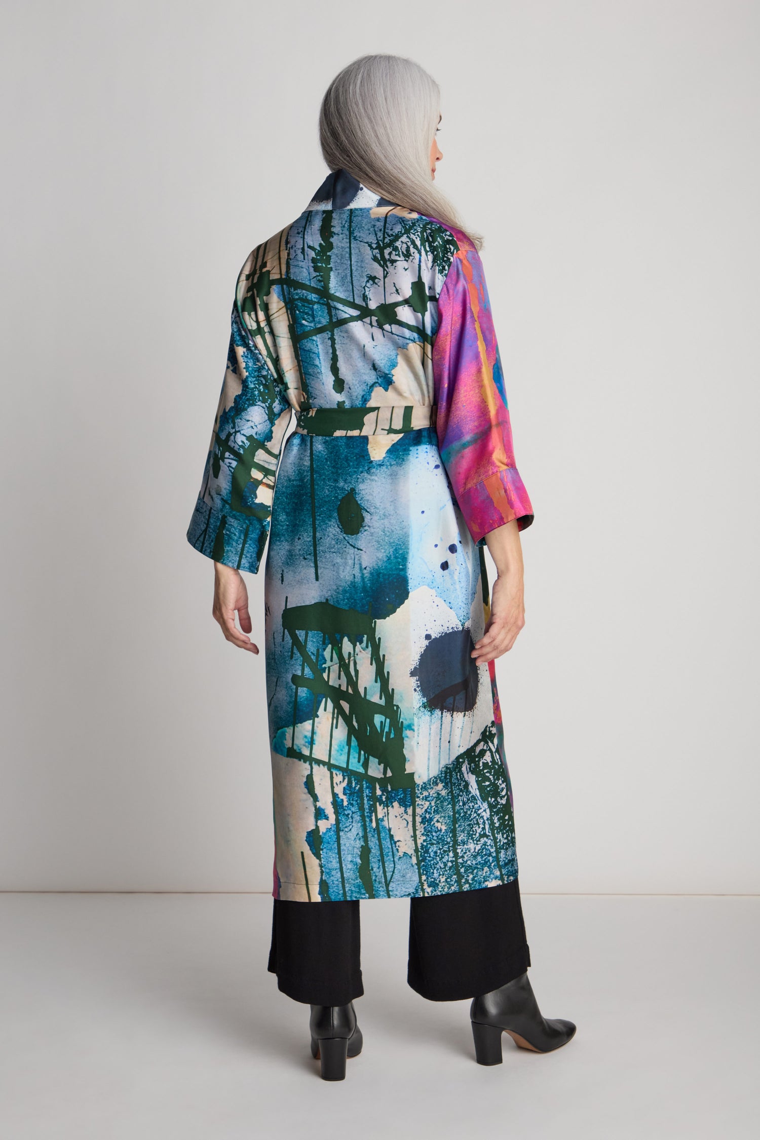 A person with long gray hair is wearing a colorful, abstract-patterned Connect the Dots Long Silk Kimono by Isabelle Gougenheim and black pants, facing away from the camera in a plain setting.