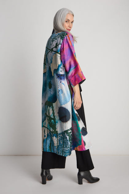 Person with long gray hair wearing a colorful, abstract-patterned long coat reminiscent of the Connect the Dots Long Silk Kimono by Isabelle Gougenheim, black pants, and black chunky heeled boots, standing against a plain light-colored background.