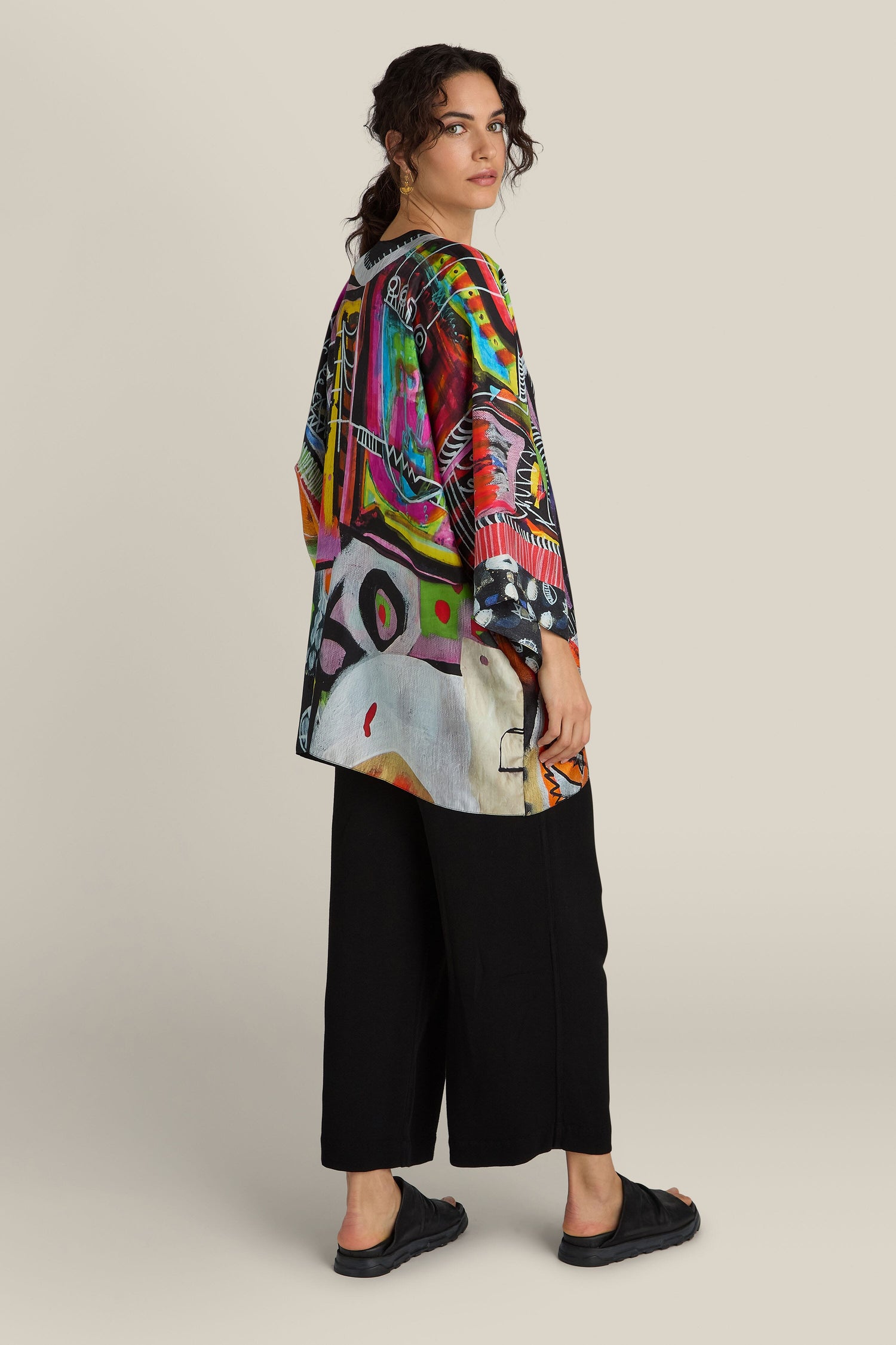 A person with curly hair is standing sideways, dressed in a vibrant, oversized top featuring artistic patterns similar to the Havana Print Short Silk Kimono by Isabelle Gougenheim. They have paired it with loose black pants and black sandals, all set against a plain background.