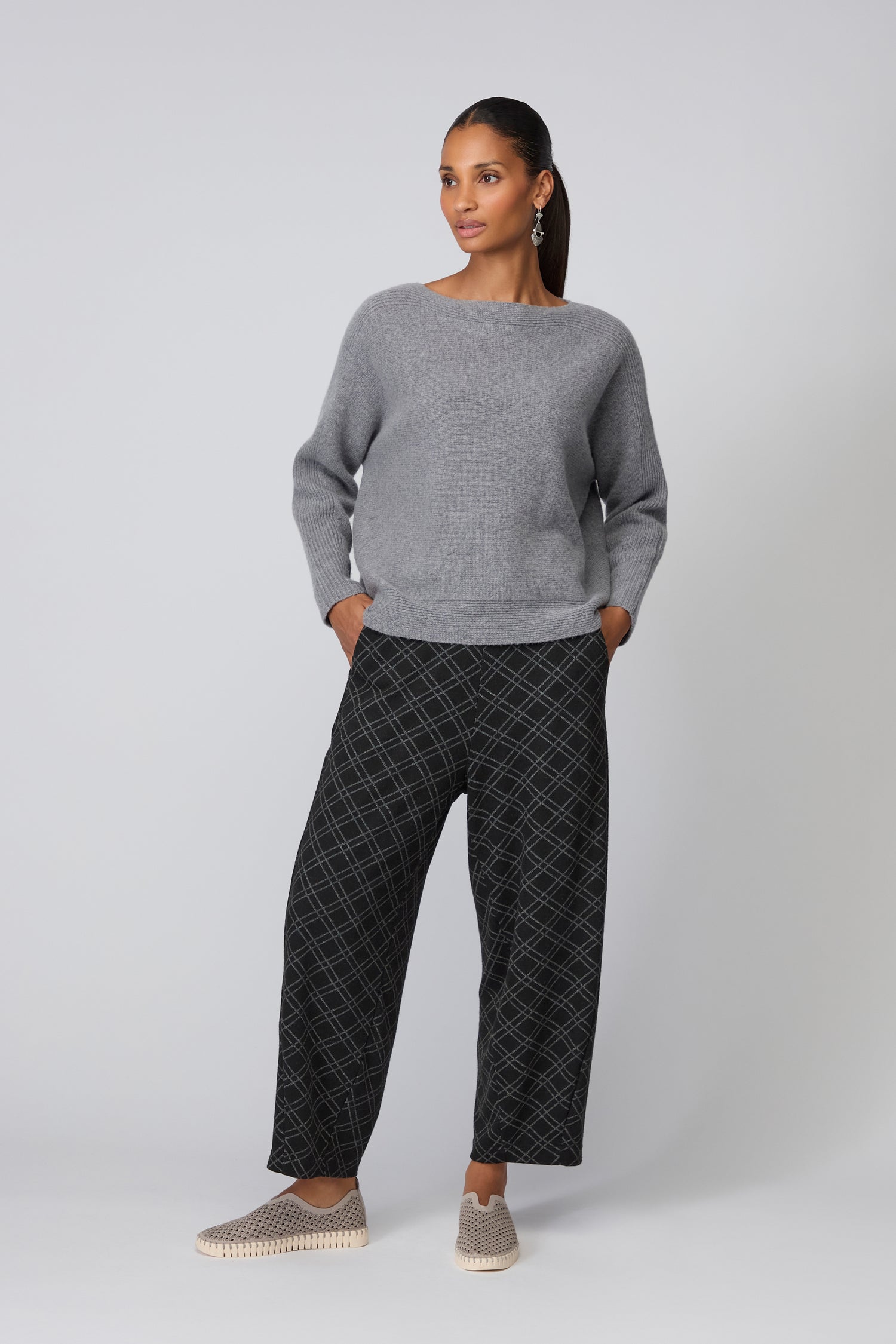 A woman stands against a plain background wearing a grey sweater, black patterned wide-leg pants, and Grey Tulip Slip On Shoes—a perfect touch of Scandinavian design.