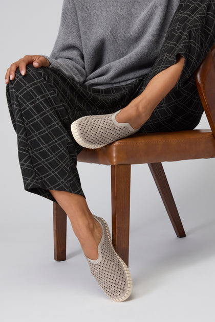 A person seated on a brown chair wearing gray plaid pants, a gray sweater, and Grey Tulip Slip On Shoes. Only the lower body is visible, highlighting sustainable shoes inspired by Scandinavian design.