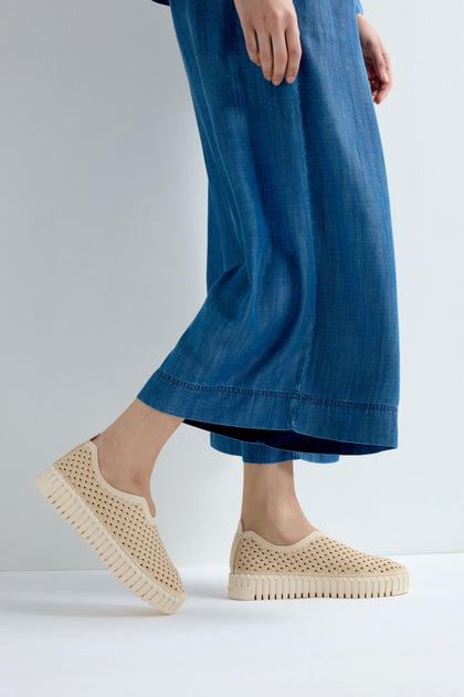 A person wearing a long blue denim skirt and beige Tulip Slip On Shoes with a perforated design and platform sole stands against a plain background.
