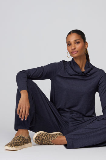 A woman sits on the floor against a light background, wearing a dark long-sleeve lounge set with wide-leg pants, gold hoop earrings, and Tulip Leopard Slip On Shoes.