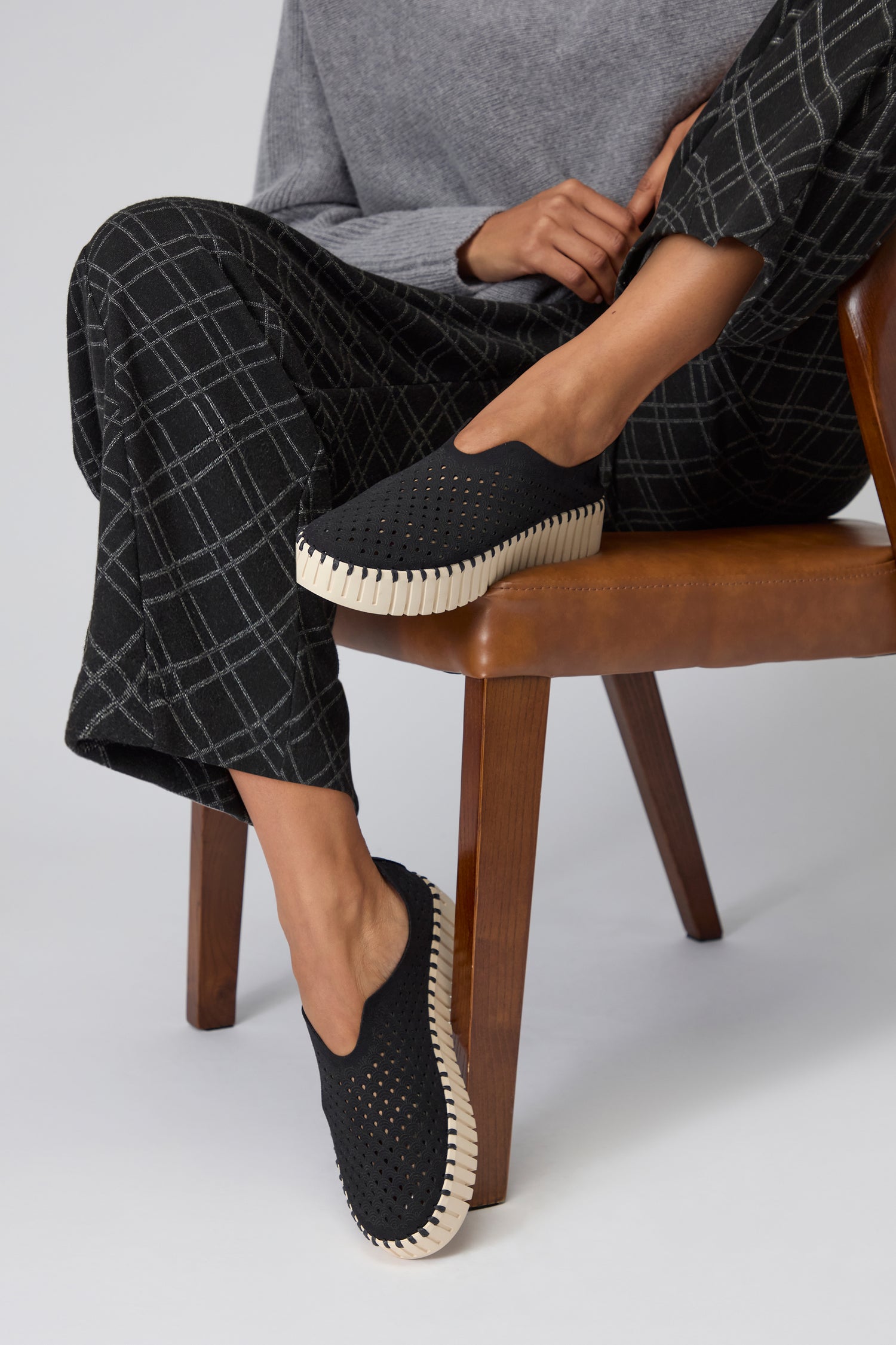 A person sitting on a wooden chair wears Black Tulip Platform Slip On Shoes with white soles and black plaid pants, showcasing stylish sustainable footwear.