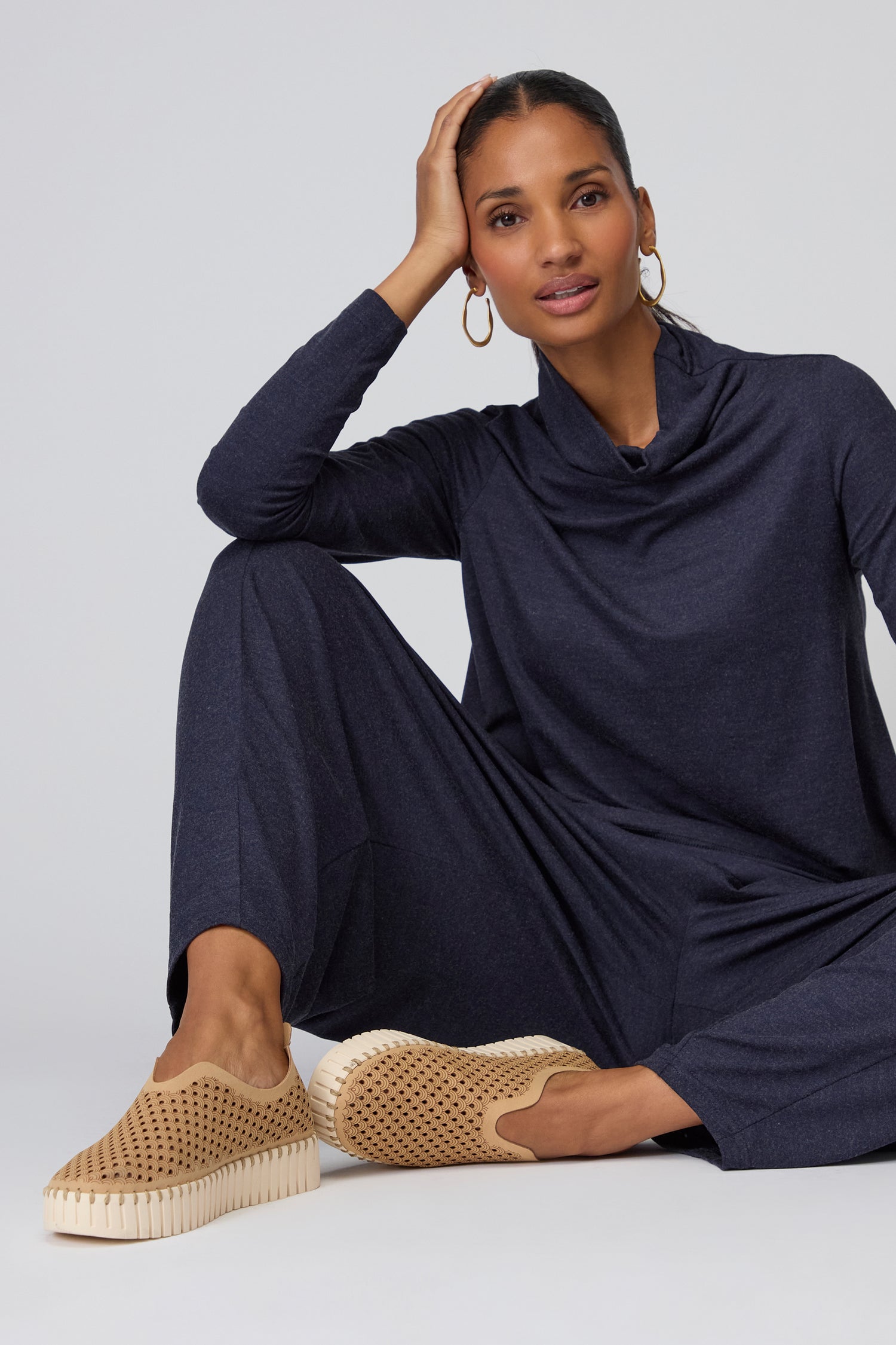 A woman sits on the floor with one knee up, wearing a navy long-sleeve outfit and Latte Tulip Platform Slip On Shoes, featuring a recycled microfibre upper, looking at the camera with her hand resting on her head.