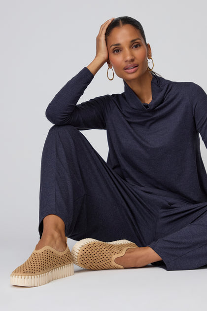 A woman sits on the floor with one knee up, wearing a navy long-sleeve outfit and Latte Tulip Platform Slip On Shoes, featuring a recycled microfibre upper, looking at the camera with her hand resting on her head.