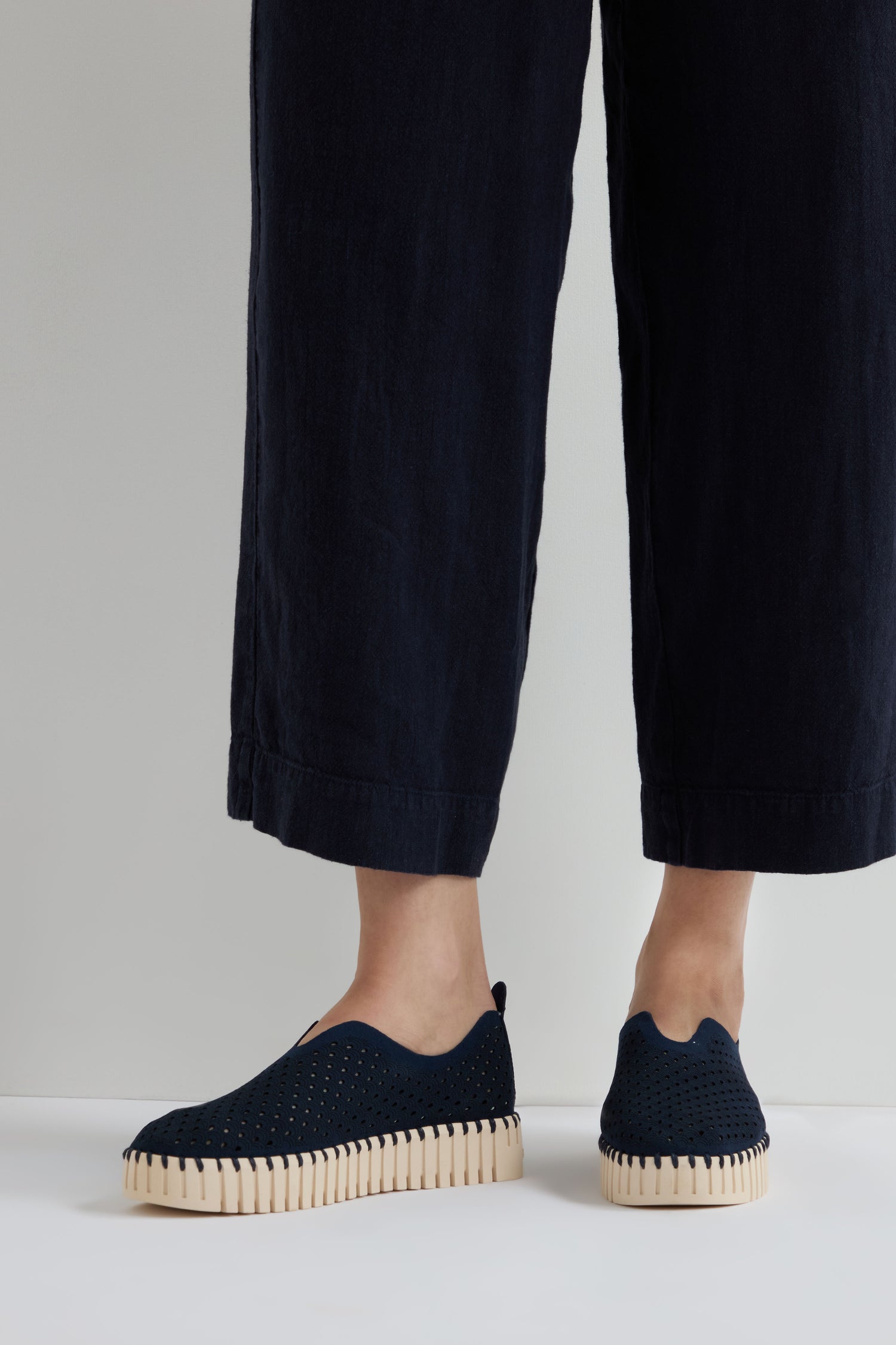 Person wearing indigo Tulip Platform slip-on shoes, styled with black wide-leg pants and standing against a plain background.