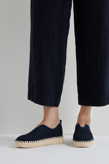 Person wearing indigo Tulip Platform slip-on shoes, styled with black wide-leg pants and standing against a plain background.