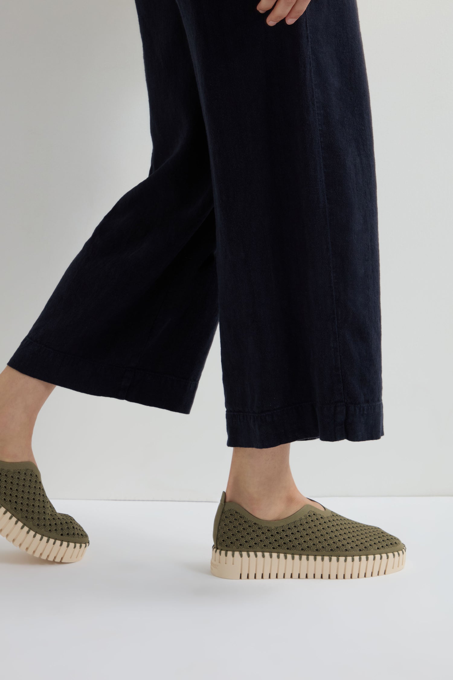 A person wearing wide-leg black pants and Khaki Tulip Platform Slip On Shoes with a perforated design and beige soles, walking on a white surface.