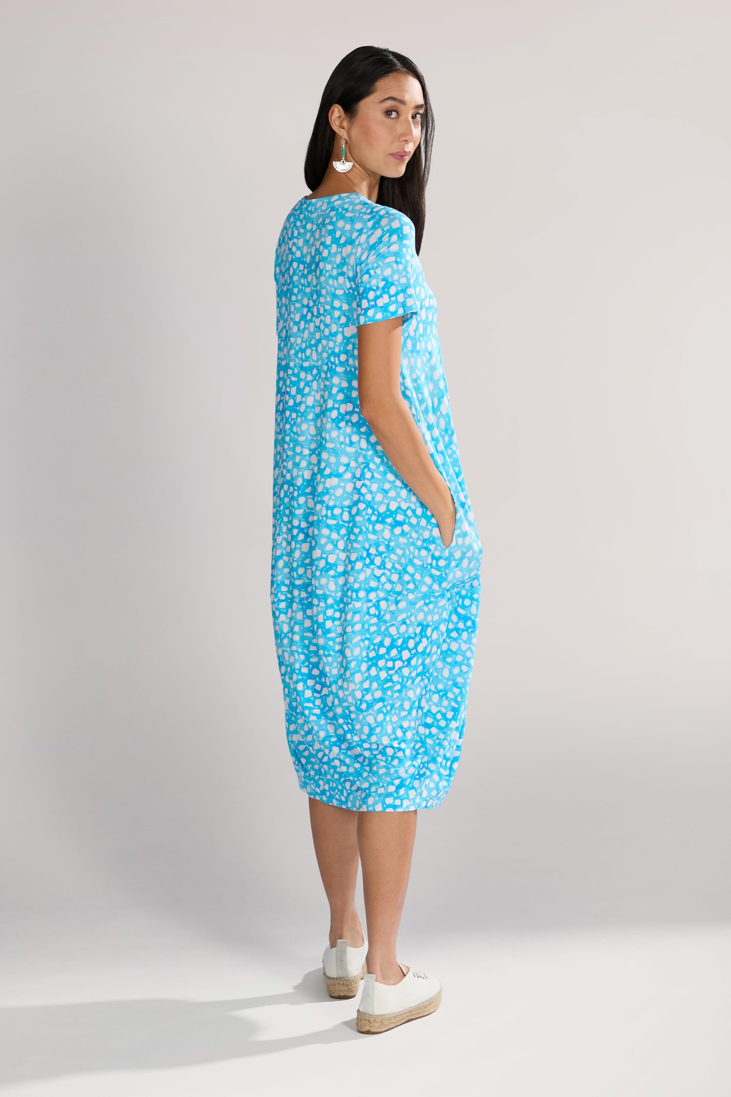 A woman with long dark hair wears a blue Pebble Jersey Print Bubble Dress featuring an abstract bubble motif and pockets. With white shoes completing her midi-length look, she stands against a neutral background, looking over her shoulder.