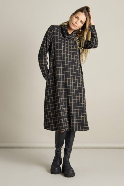 Against a plain background, an individual stands wearing the Broken Check Jersey Dress, featuring a cosy funnel cowl neck. The knee-length dress with long sleeves is paired with black leggings and black boots for a contemporary twist. They have one hand comfortably tucked into a pocket.