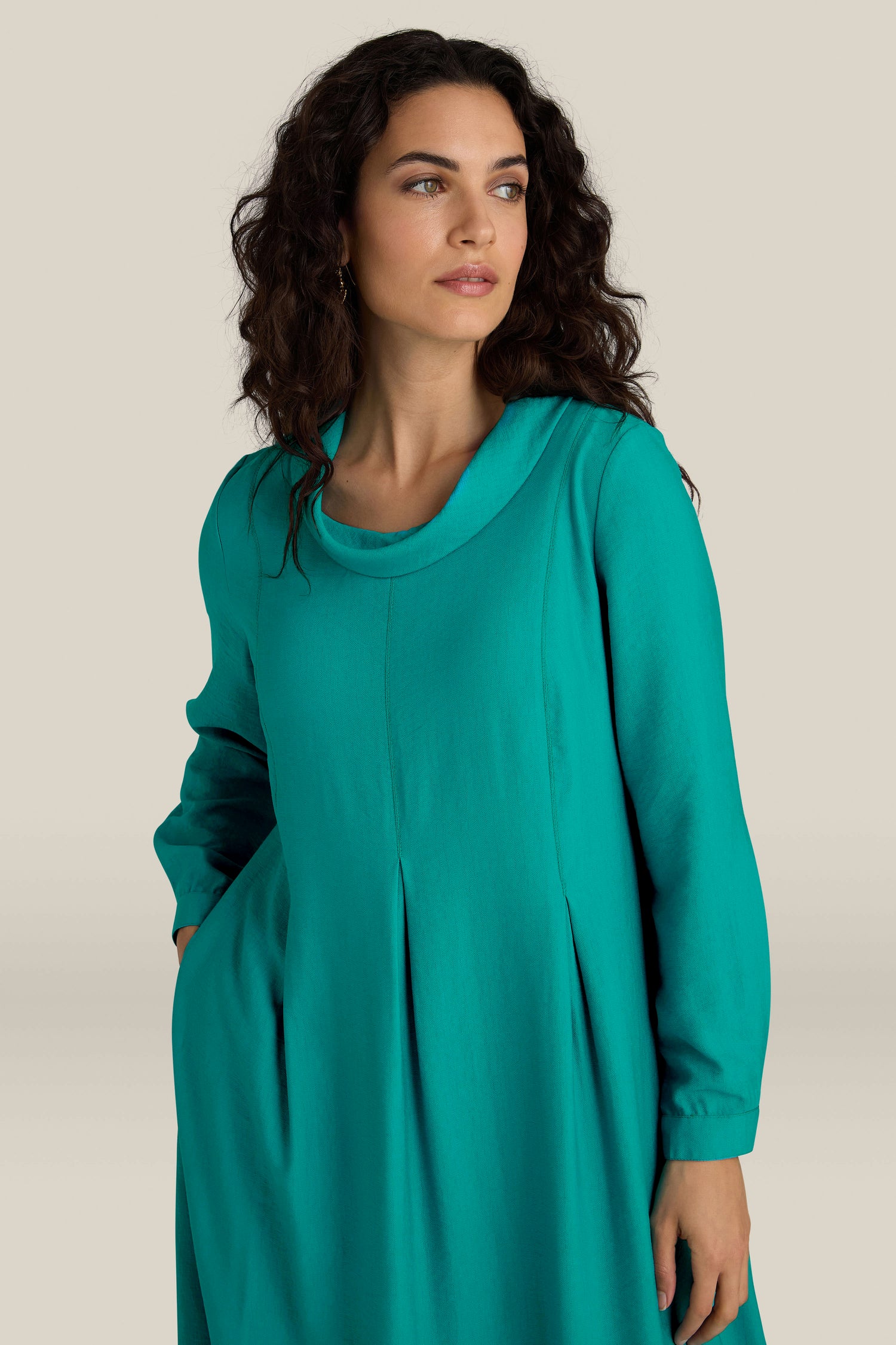 A woman with curly hair, epitomizing everyday elegance, stands against a plain background wearing a long-sleeve teal Textured Viscose Bubble Dress, looking to the side.