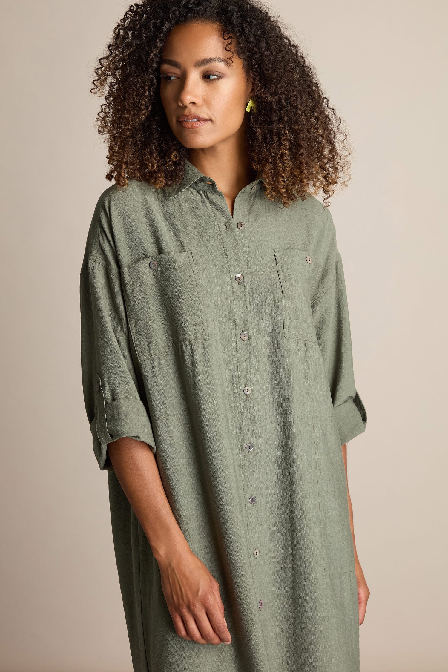 A woman with curly hair wears a green Textured Viscose Shirt Dress made of mid-weight fabric. She looks slightly to the side, with one hand relaxed by her side.