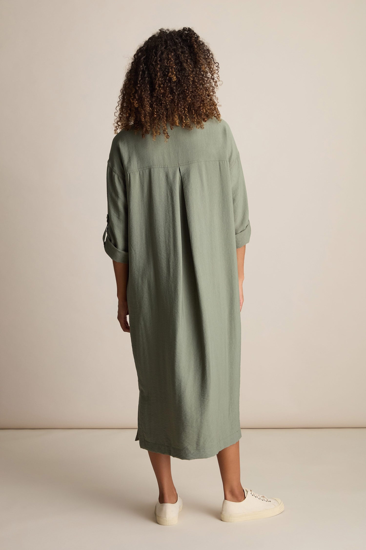 A person with curly hair is standing and facing away, wearing a loose-fitting, knee-length Textured Viscose Shirt Dress in green and white shoes against a plain background.