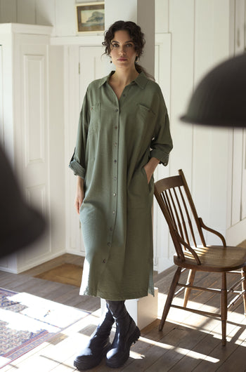 A woman in the Textured Viscose Shirt Dress, showcasing its timeless khaki colourway and mid-weight silky textured fabric, stands by a wooden chair in a well-lit room with white walls and wooden floors.