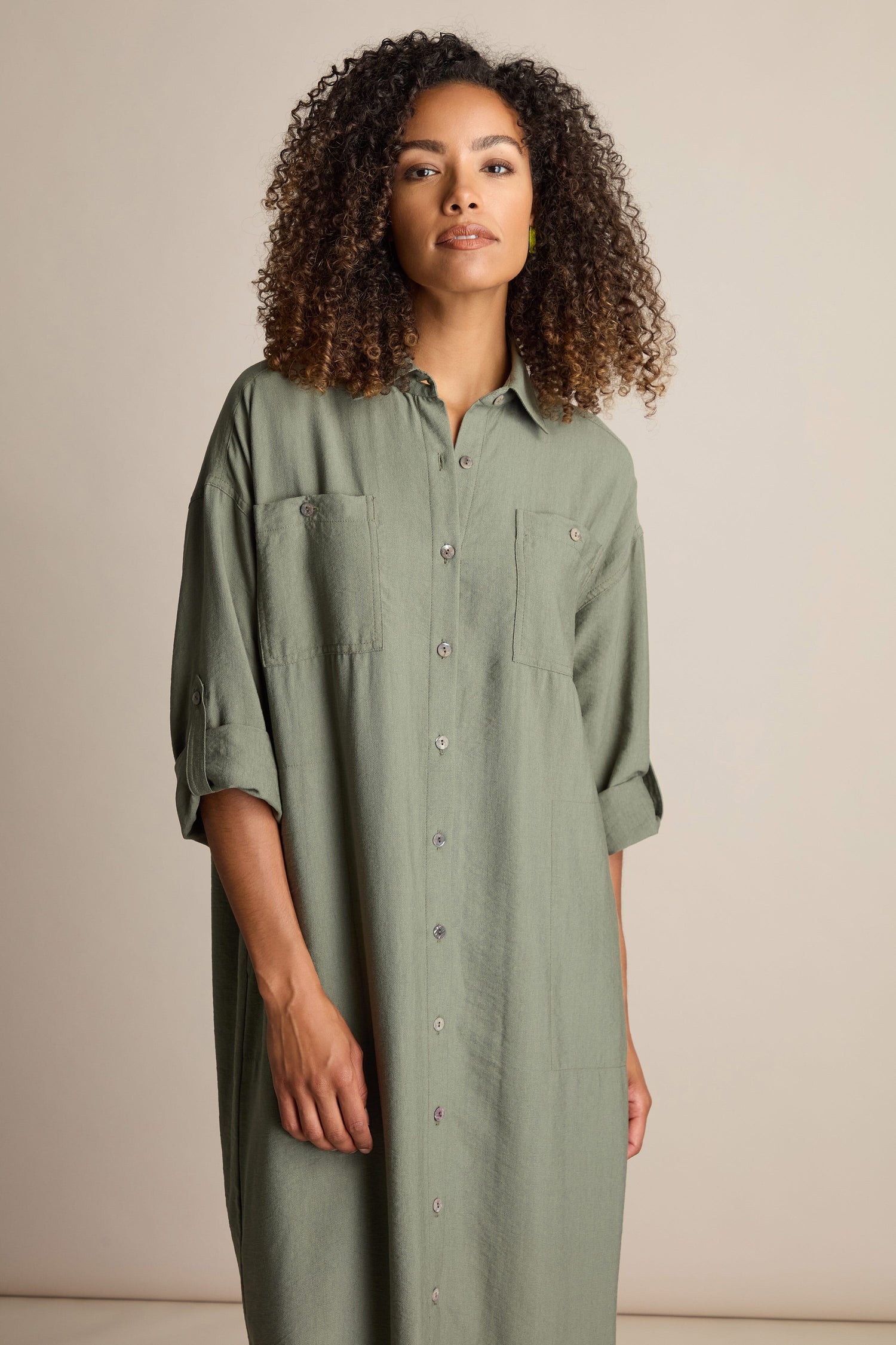 A person with curly hair wears the Textured Viscose Shirt Dress, which features a loose fit and rolled-up sleeves in a long green classic design, standing against a plain background to highlight the khaki colourway of the mid-weight viscose fabric.