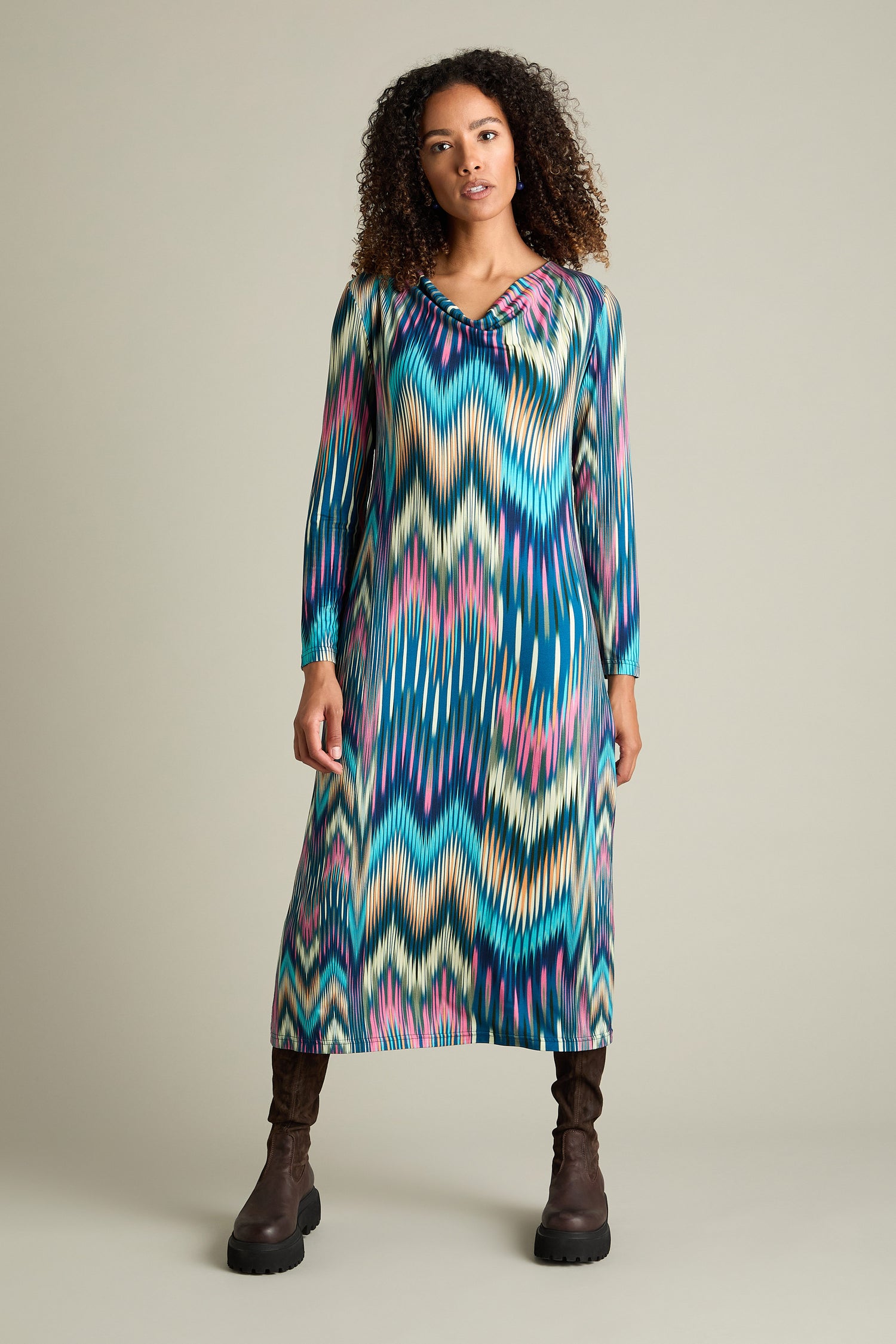 A person wearing the Autumnal Ikat Jersey Dress, which features a long, colorful pattern and a cowl-neck design, stands facing forward with their arms relaxed by their sides. They have curly hair and are also wearing tall, dark boots.