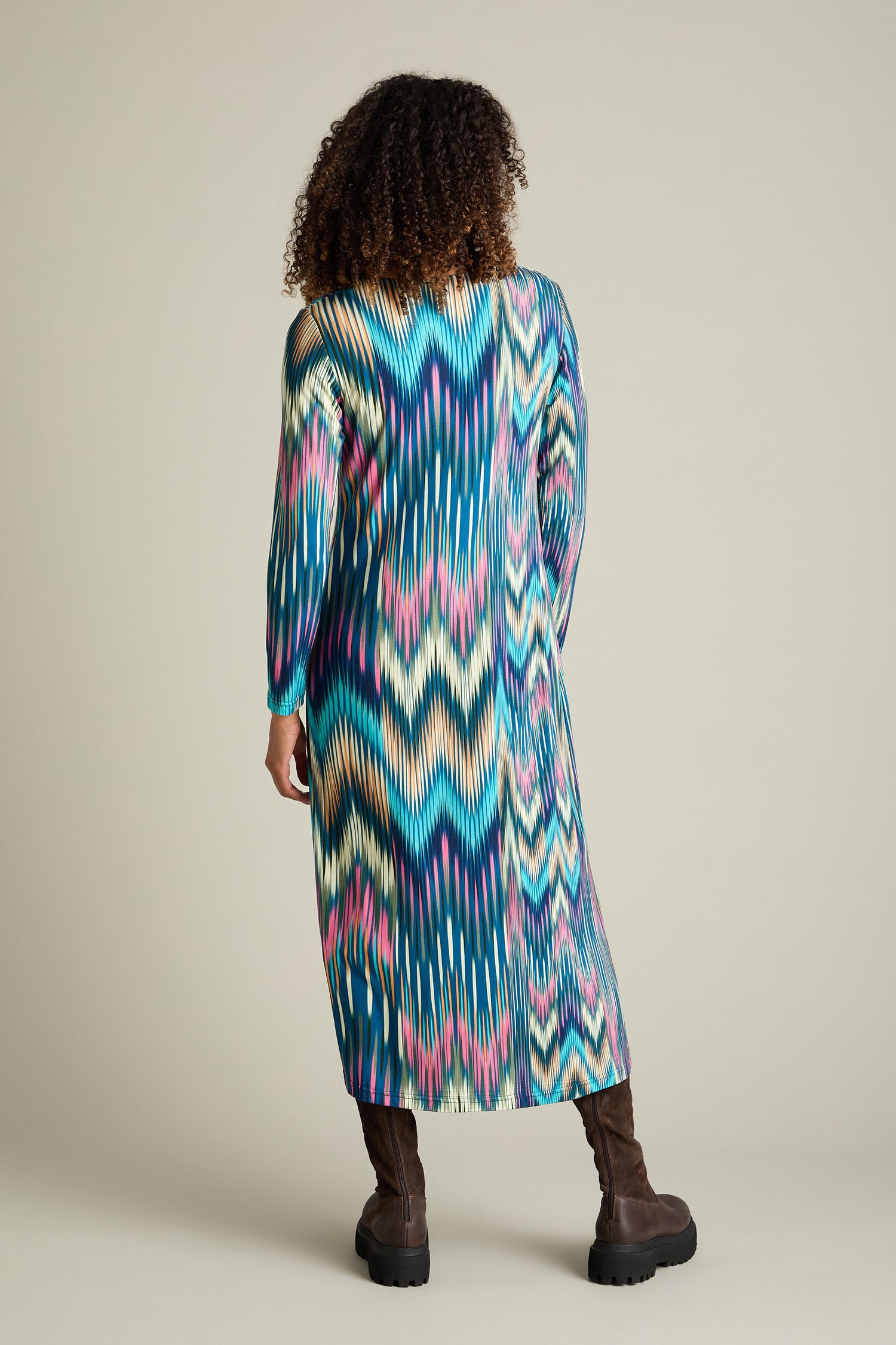 Person standing with back to camera, wearing an Autumnal Ikat Jersey Dress featuring a long, colorful zigzag-patterned print and brown boots.