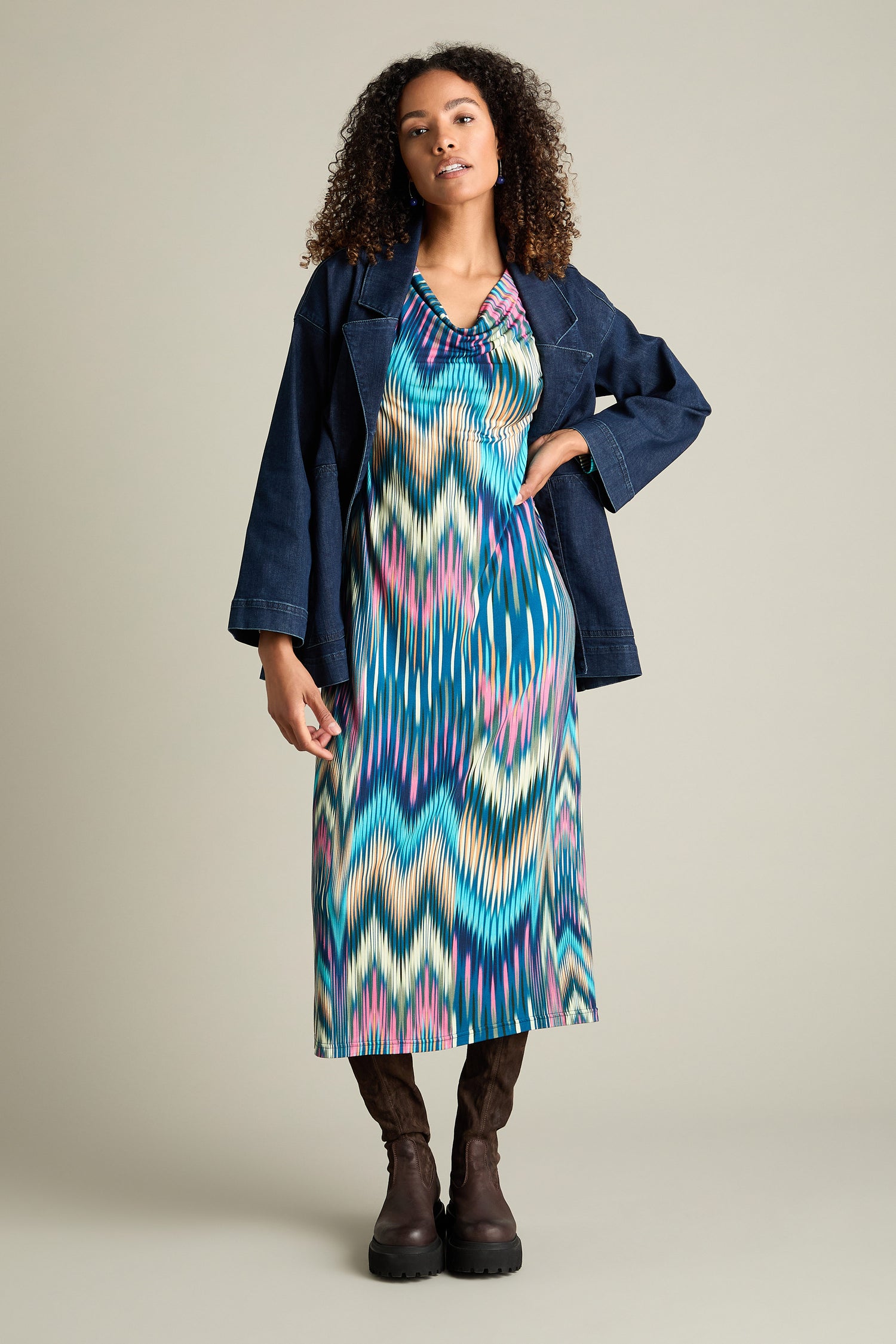 A person with curly hair is wearing an Autumnal Ikat Jersey Dress, a denim jacket, and boots. The person is standing in front of a plain background.