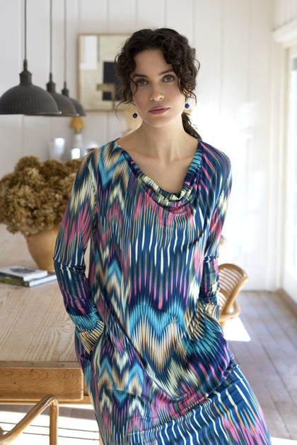 A person with curly hair wearing an Autumnal Ikat Jersey Dress stands in a modern, well-lit room with a wooden table and chairs in the background.