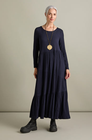 A woman with grey hair is wearing a Crinkle Viscose Tiered Dress, navy blue in color and maxi length, paired with black boots. The dress features an elegant A-line silhouette. She sports a long necklace with a large circular pendant and stands against a plain background.