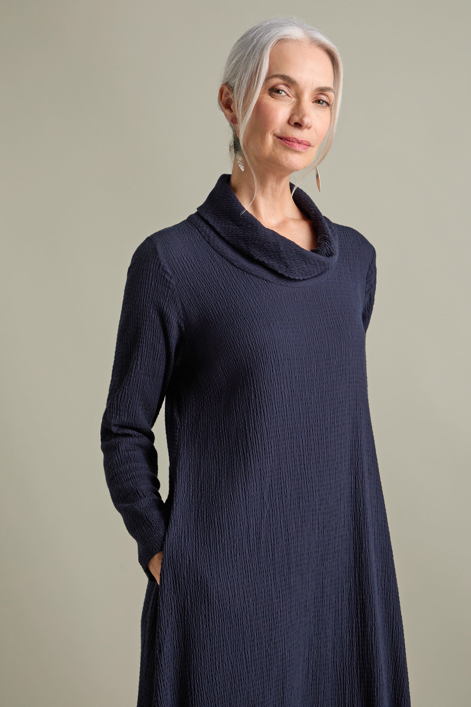 An older woman with gray hair stands with her hands in her pockets, donning a dark, long-sleeve Crinkle Viscose Bubble Dress. She gazes at the camera with a relaxed, chic demeanor, showcasing the dress's voluminous silhouette.