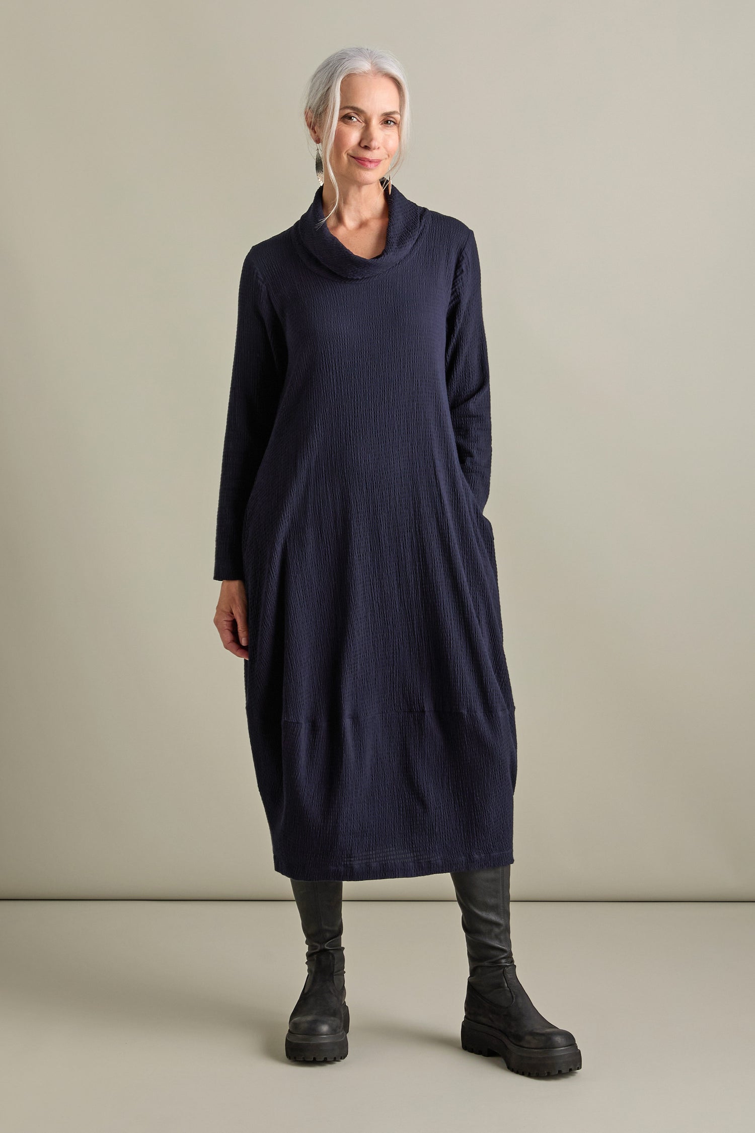 A woman with white hair, dressed in a voluminous dark blue Crinkle Viscose Bubble Dress and black boots, stands against a plain background, epitomizing relaxed chic.