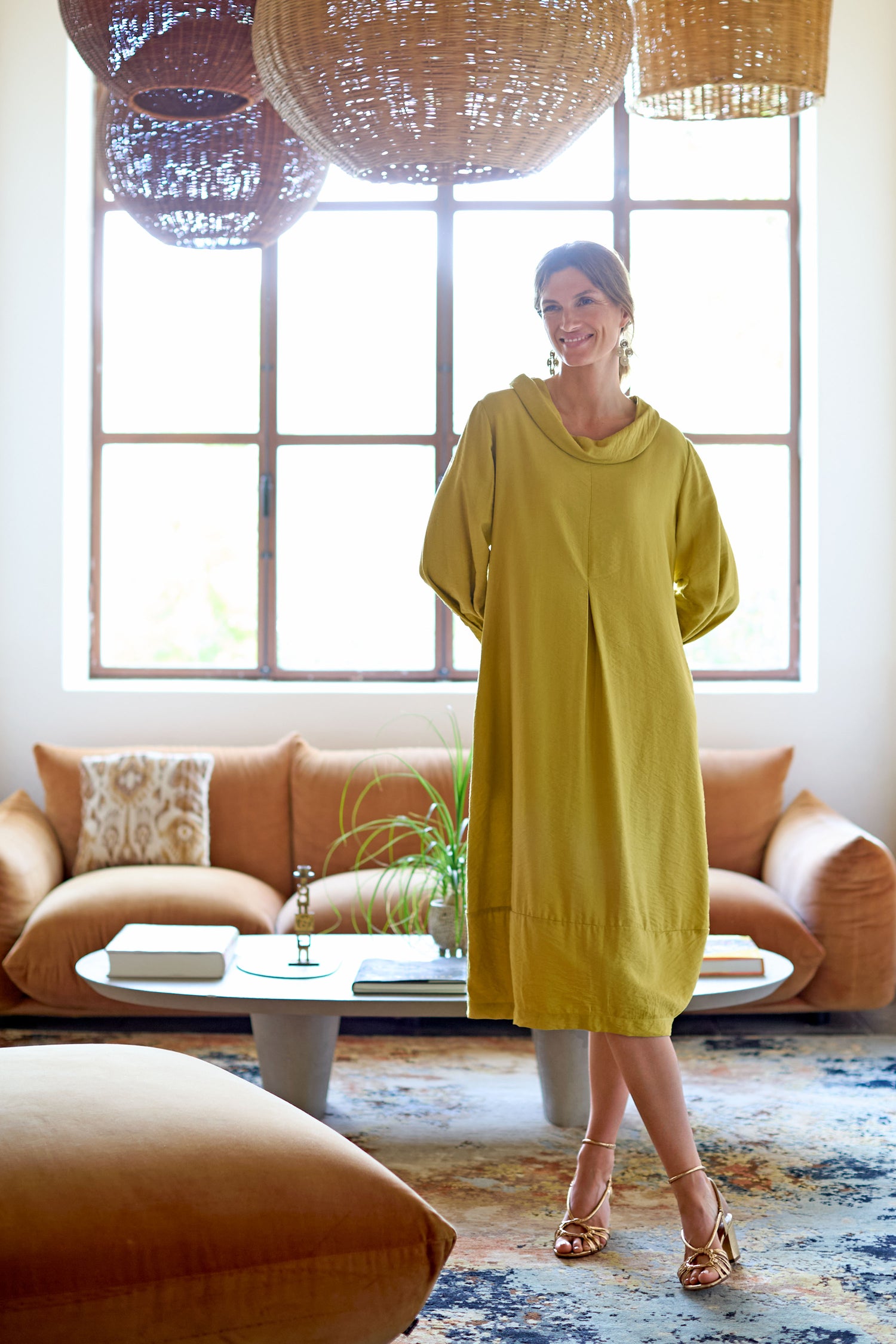 Wearing a Textured Viscose Bubble Dress, someone stands in a chic living room featuring a brown sofa, round coffee table, and large woven light fixtures. The dress's flattering fit perfectly complements the contemporary elegance of the space.