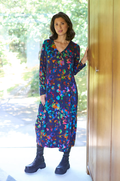 A woman stands indoors by a wooden wall, wearing the Vivid Petals Jersey Dress—a long-sleeved, colorful floral dress—paired with black boots. Trees can be seen outside through the large window beside her.