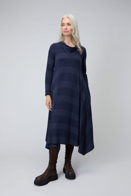 A woman with long gray hair models the Soft Viscose Jersey Stripe Dress in dark blue, featuring subtle stripes, an asymmetric hem, and long sleeves. She pairs it with brown knee-high boots against a plain light background.