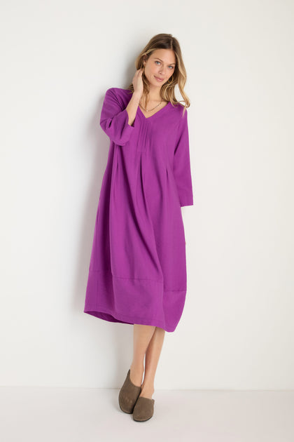 A woman stands against a white wall wearing the Textured Viscose Bubble Dress in orchid purple, a loose-fitting, long-sleeved midi dress, paired with brown slip-on shoes.