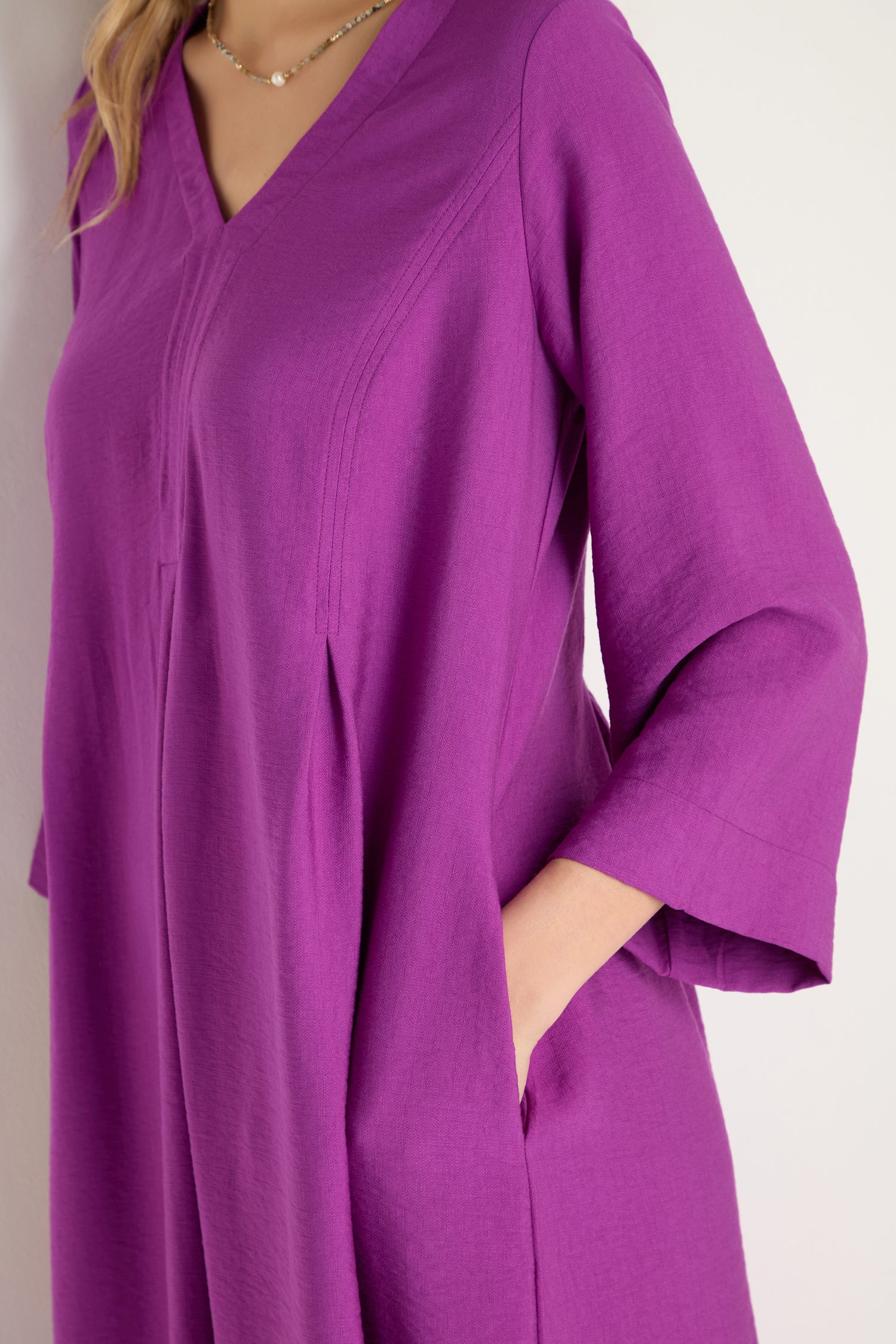 Wearing the Textured Viscose Bubble Dress in orchid purple, featuring long sleeves, a V-neckline, and side pockets, standing against a light-colored wall.